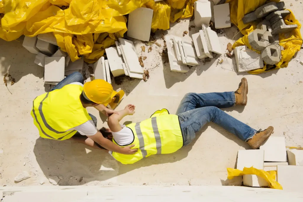 Two construction workers in safety vests attending to an injured colleague on a site with debris and yellow tarps. Two construction workers in safety vests attending to an injured colleague on a site with debris and yellow tarps.
