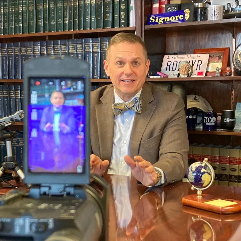 imgi_11_475457706_1196143508897975_3782572886255906941_n Lawyer in a suit speaking at a desk with a camera focused on him, bookshelves in the background.